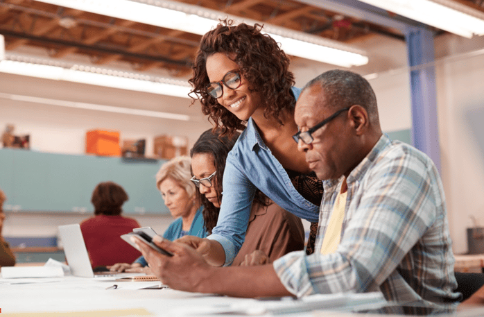 Coworkers at a table. One of them points over the shoulder at a coworker's tablet