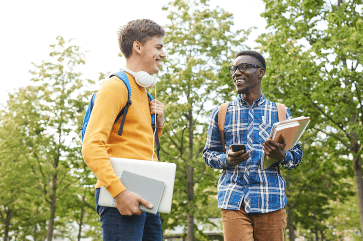 Two college students conversing outside, holding laptops and notebooks